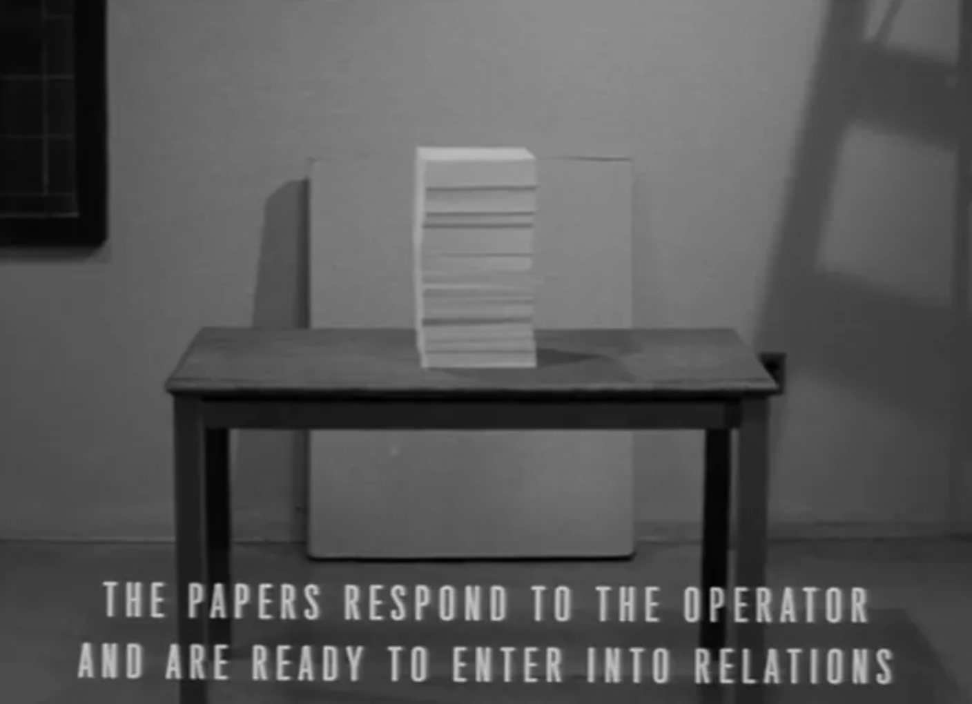 Zoe Beloff, The Infernal Dream (2011). A black and white image of a tall (2ft ish) stack of papers on an empty wooden desk in a mostly empty room. Text at the bottom reads: "THE PAPERS RESPOND TO THE OPERATOR AND ARE READY TO ENTER INTO RELATIONS."